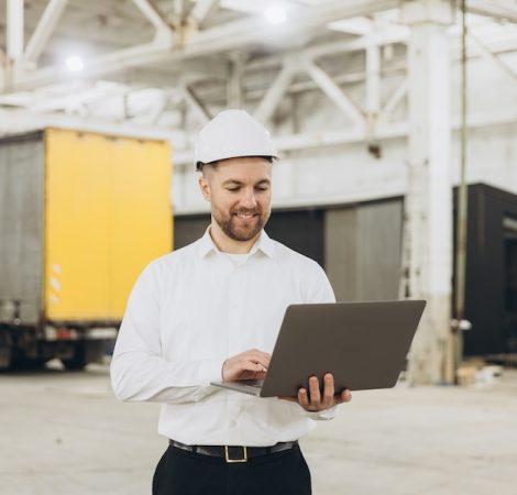 Smiling engineer wearing hardhat using laptop in modular building factory, managing production and logistics